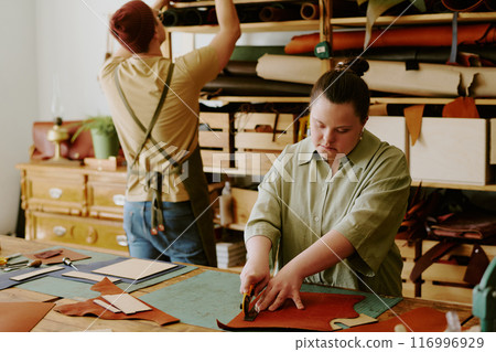 Female tailor with down syndrome cutting out leather workpieces while male tailor looking for material on wooden shelves 116996929