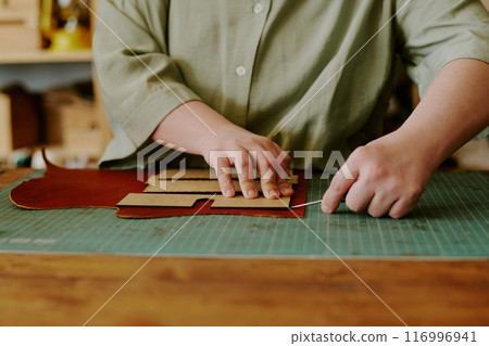 Medium close up of female hands holding knife to cut out leather details, leather piece on old wooden table in atelier 116996941