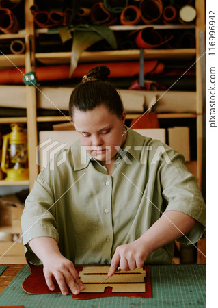 Female atelier worker with down syndrome cutting out details from leather piece very carefully 116996942