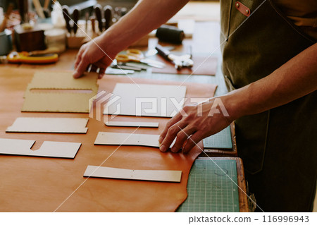 Unrecognizable male hands laying out wooden shapes on milk chocolate colored leather piece to cut out future details 116996943