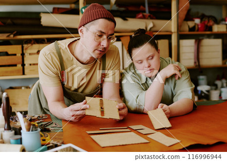 Young male tailor holding wooden shape while discussing future good with his female intern with down syndrome 116996944