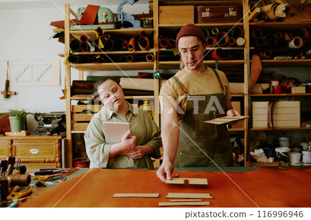 Male tailor choosing wooden shape to create leather good while his female assistant with down syndrome watching process and holding notebook Male tailor choosing wooden shape to create leather good while his female assistant with down syndrome watching process and holding notebook 116996946