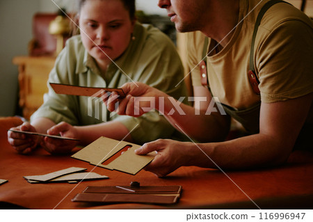 Male tailor demonstrating wooden rectangular shape to his female coworker with down syndrome while they leaning on wooden table covered with leather Male tailor demonstrating wooden rectangular shape to his female coworker with down syndrome while they leaning on wooden table covered with leather 116996947