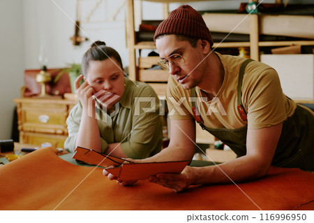 Tailor and client with down syndrome leaning on old table while examining closely leather workpieces Tailor and client with down syndrome leaning on old table while examining closely leather workpieces 116996950