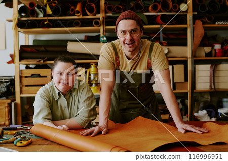 Portrait of smiling male tailor and his female client with down syndrome leaning on wooden table covered with leather workpieces while in atelier 116996951