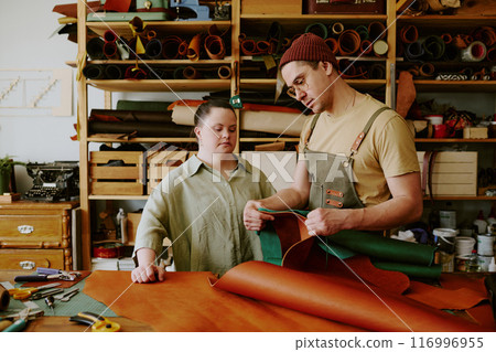 Caucasian male tailor taking measurements while his female client with down syndrome watching process, wooden shelves full of leather rolls and other materials behind them Caucasian male tailor taking measurements while his female client with down syndrome watching process, wooden shelves full of leather rolls and other materials behind them 116996955