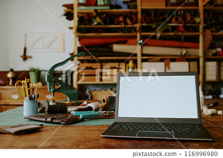 Black laptop with white screen and leather punch machine with its tool in background on old wooden table 116996980