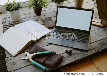 Black laptop surrounded by notes and leather goods on gray wooden table, four clay pots with plants in background 116996982