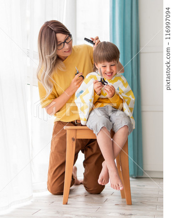 Mother makes haircut to her son at home. Little boy sits covered with cloth and holds pair of scissors. Self care at home. Happy lifestyle. Mother makes haircut to her son at home. Little boy sits covered with cloth and holds pair of scissors. Self care at home. Happy lifestyle. 116997094