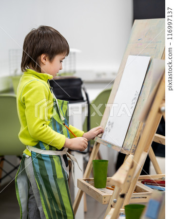Little boy draws in studio. Art therapy, art education for children. Painting on paper on easel. Developing fine motor skills and imagination. 116997137