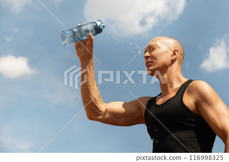 Sweaty handsome sporty man refreshing himself water from bottle against blue sky at hot summer after training outdoors. Concept water balance. Close up. Banner. Sweaty handsome sporty man refreshing himself water from bottle against blue sky at hot summer after training outdoors. Concept water balance. Close up. Banner. 116998325