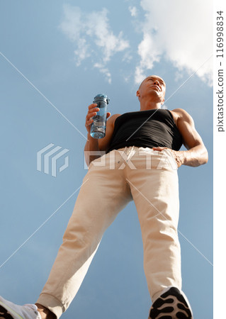 Athletes handsome sun-tanned man drinking water from bottle against sky. Concept water balance for hot season for active workout. Hero view. Vertical. 116998834