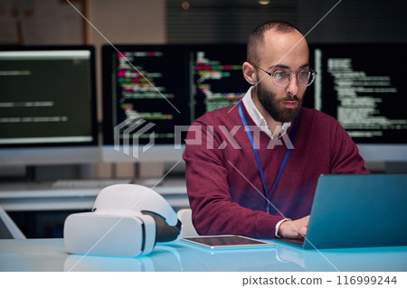 Portrait of bearded adult man wearing glasses and using laptop at desk in IT development office , copy space 116999244