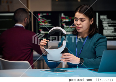 Portrait of young woman setting up VR headset while developing new technology in office with code screens in background, copy space 116999245