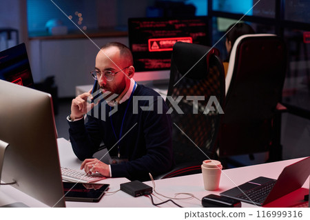 High angle view at bearded adult man using computer with multiple monitors in cybersecurity department and speaking on phone High angle view at bearded adult man using computer with multiple monitors in cybersecurity department and speaking on phone 116999316