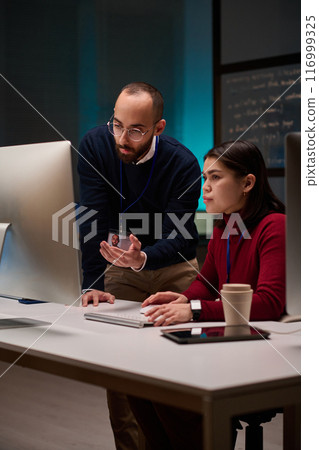 Vertical portrait of cybersecurity team of two people using computer together in office lit by blue light Vertical portrait of cybersecurity team of two people using computer together in office lit by blue light 116999325