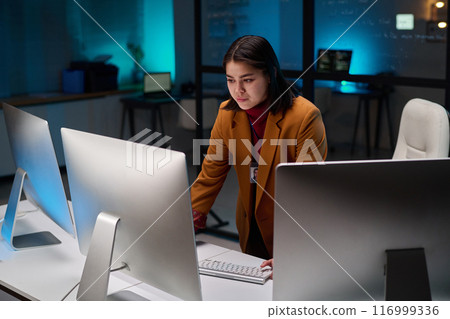 Portrait of female cybersecurity expert using computer with multiple monitors in office lit by blue light 116999336