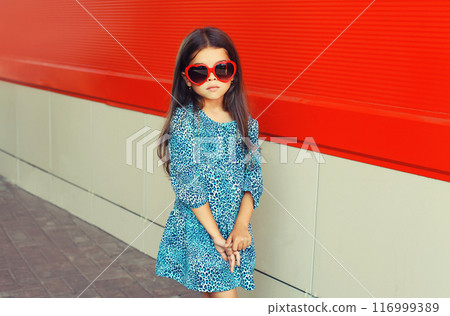 Beautiful little girl child in red heart shaped sunglasses and blue dress on city street 116999389