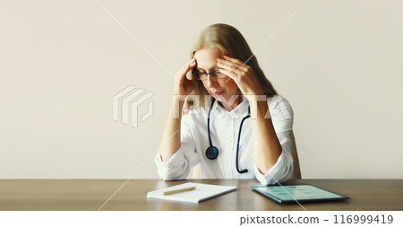 Tired woman doctor working with documents while sitting at desk in medical clinic office 116999419