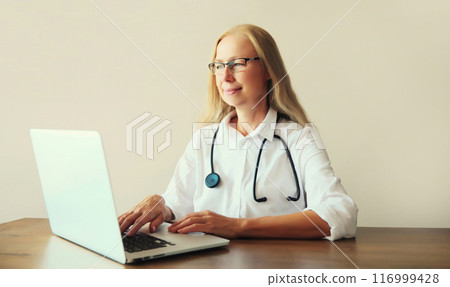 Happy woman doctor working with laptop computer while sitting at desk in medical clinic office 116999428