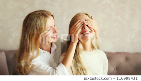 Happy smiling caucasian adult daughter covering mother's eyes making surprise at home Happy smiling caucasian adult daughter covering mother's eyes making surprise at home 116999431