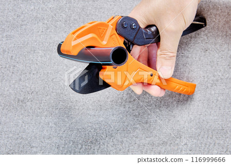 Piece of plastic pipe lies between blades of pipe cutter with ratcheting mechanism, plumber hand holds tool. Piece of plastic pipe lies between blades of pipe cutter with ratcheting mechanism, plumber hand holds tool. 116999666