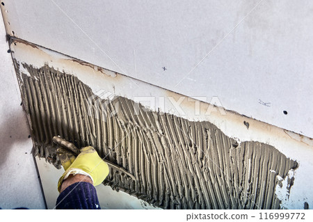 Builder tiler applies adhesive cement mortar to wall using comb trowel. Builder tiler applies adhesive cement mortar to wall using comb trowel. 116999772