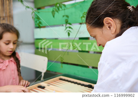 Mother and daughter playing backgammon together in a cozy outdoor garden setting 116999902
