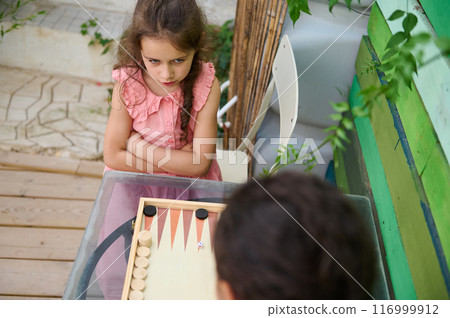 Young girl playing backgammon with friend outdoors, expression of concentration Young girl playing backgammon with friend outdoors, expression of concentration 116999912