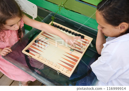 Mother and daughter playing an engaging game of backgammon on a glass table outdoors Mother and daughter playing an engaging game of backgammon on a glass table outdoors 116999914