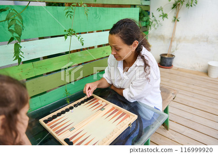 Woman playing backgammon with child outdoors on wooden deck with green painted backdrop Woman playing backgammon with child outdoors on wooden deck with green painted backdrop 116999920