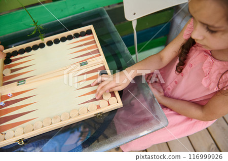 Young girl playing backgammon outdoors on a glass table wearing a pink dress 116999926