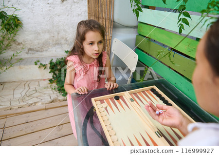 Young girl playing backgammon outdoors with adult, learning and focused expression 116999927