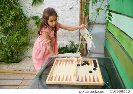 Young girl in pink dress playing backgammon outdoors in a garden setting Young girl in pink dress playing backgammon outdoors in a garden setting 116999960