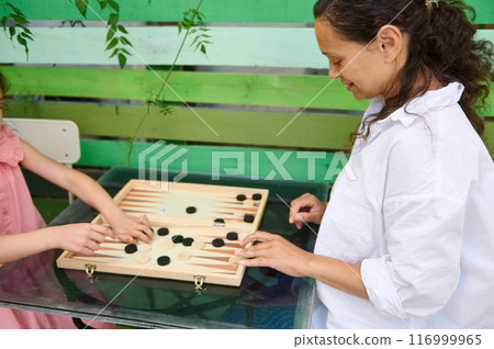Mother and daughter playing backgammon outdoors on a glass table 116999965