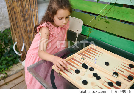 Young girl playing backgammon at home. Cute child in pink dress focused on game indoors Young girl playing backgammon at home. Cute child in pink dress focused on game indoors 116999966