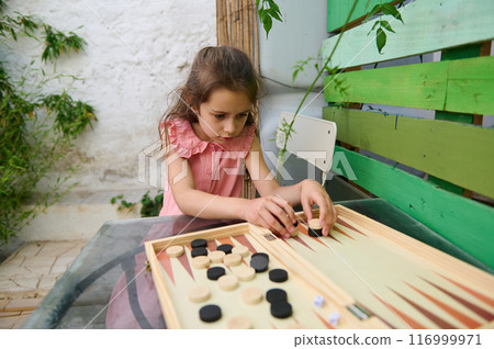 Focused young girl playing backgammon game outdoors on a sunny day 116999971
