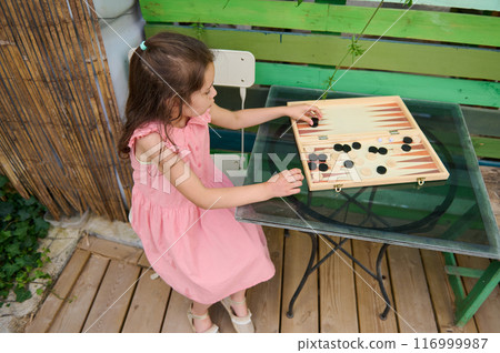 Little girl in pink dress playing backgammon outside on a wooden deck 116999987