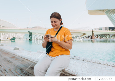 Young traveling woman using phone outdoors. Concept of travel, tourism and vacation in city. Ciudad de las Artes y las Ciencias de Valencia.Valencia City of Arts and Sciences. High quality photo 117000972