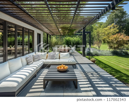 A modern patio with a pergola, white sectional sofa, coffee table with fruit bowl, and dining area, overlooking a lush garden. 117002343