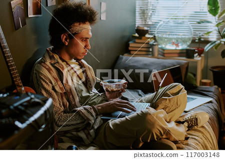 University student checking his social networks and eating Chinese food while chilling on bed 117003148