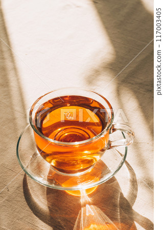 A cup of tea in glass cup and saucer illuminated by warm sunlight on a light table with a simple tablecloth. Vertical frame, copy space 117003405