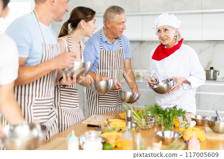 Smiling old woman chef of cooking course teaching attendees how to mix sauce Smiling old woman chef of cooking course teaching attendees how to mix sauce 117005609