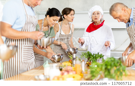 Smiling old woman chef of cooking course teaching attendees how to mix sauce 117005712