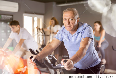 Portrait of mature man taking indoor cycling class at fitness center, doing cardio riding bike Portrait of mature man taking indoor cycling class at fitness center, doing cardio riding bike 117005732
