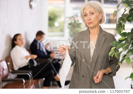 Unsatisfied displeased mature woman frowns face stands in hall of directors reception room 117005797