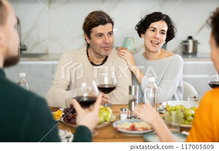 Group of joyful individuals are celebrating a holiday, enjoying drinks and conversation around table in their home 117005809