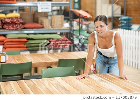Young girl thoughtfully evaluating wooden patio table in garden store 117005879