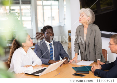 Senior woman head of department speaks at meeting and reports unpleasant news to frowning colleagues 117005941