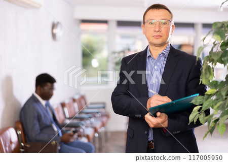 Adult man in glasses with documents stands in reception 117005950